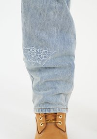 Světle modré džíny z denimu s ohrnutým lemem, s bílým vyšitým logem na nohavici. Doplňuje je hnědá šněrovací kotníková bota se zlatými očky.
