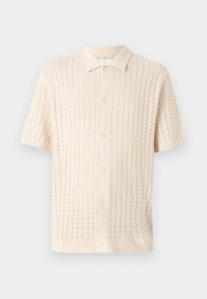 Chemise à manches courtes à boutons en crème clair, présentant un motif tricoté texturé avec un col et cinq boutons sur le devant.
