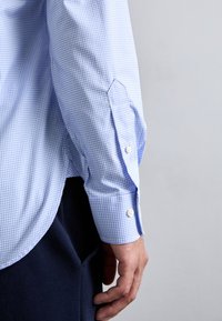 Camicia a quadri blu e bianca con maniche lunghe, dotata di bottoni bianchi e una vestibilità su misura. La fine della manica presenta un design a polsino leggermente appuntito.