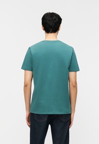 Άνδρας με κοντά μαύρα μαλλιά, φορώντας μια τιρκουάζ κοντομάνικη μπλούζα και σκούρο μπλε τζιν, στέκεται γυρισμένος με την πλάτη προς το λευκό φόντο.