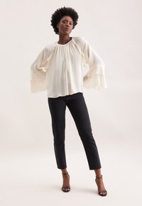 Blusa color crema con maniche a campana con volants e scollatura arricciata, abbinata a jeans neri slim fit e sandali neri con tacco a strappo.