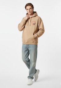 Felpa beige con cappuccio e grafica a forma di cuore rosso, abbinata a jeans azzurri chiari distressed e sneakers bianche, in piedi su uno sfondo neutro.