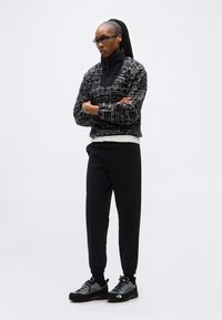 Γυναίκα που φοράει γυαλιά ηλίου, ένα μαύρο και άσπρο σακάκι με σχέδια, μαύρο παντελόνι και γκρι παπούτσια πεζοπορίας, στέκεται με τα χέρια σταυρωμένα σε λευκό φόντο.