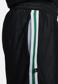 Pantalones deportivos negros con un bolsillo con cremallera, tejido texturizado y franjas laterales en blanco y verde. Destacados con una pequeña etiqueta de logo.