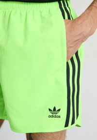 Shorts sportive de culoare verde neon, cu talie ajustabilă prin șnur, buzunare laterale și accente cu trei dungi negre. Prezintă un logo brodat negru.