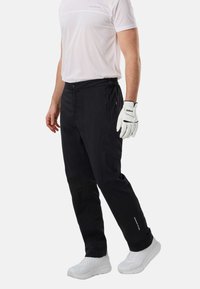 Homme portant un pantalon de golf noir, un T-shirt blanc, un gant de golf blanc et des chaussures de sport blanches, debout sur fond blanc.