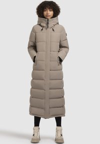 Abrigo largo de plumas color beige claro con capucha, que presenta paneles acolchados, cierre de cremallera y bolsillos laterales. Se lleva con leggings negros y botas beige.