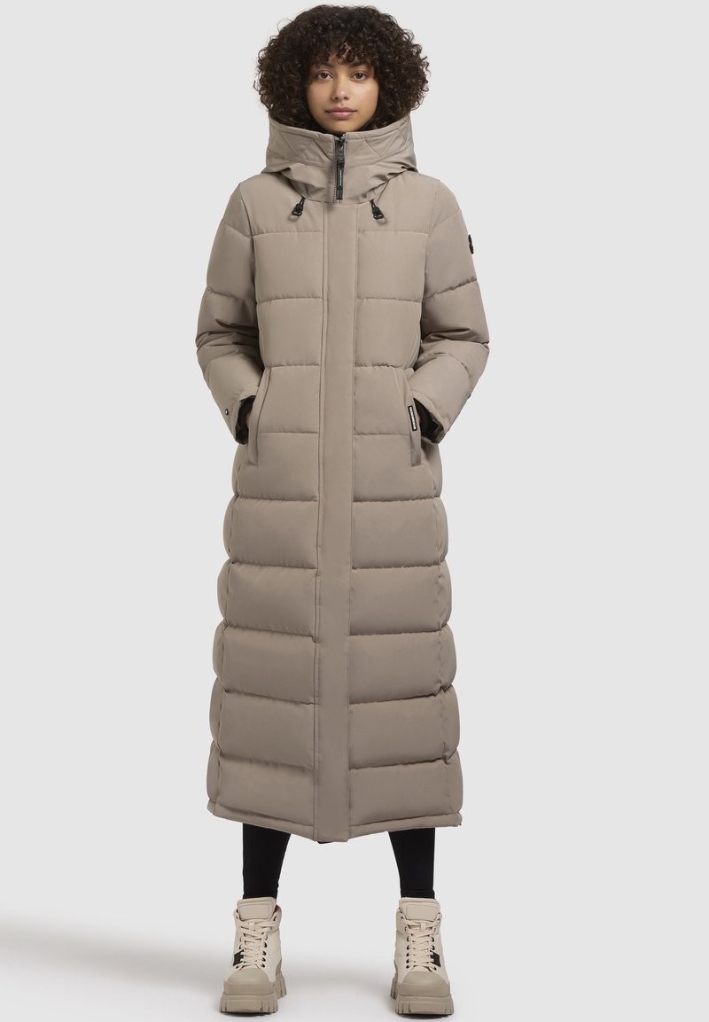 Abrigo largo de plumas color beige claro con capucha, que presenta paneles acolchados, cierre de cremallera y bolsillos laterales. Se lleva con leggings negros y botas beige.