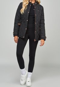 Chaqueta acolchada negra con herrajes en dorado rosa, cuello alto y bolsillos con cremallera; combinada con leggings negros y zapatillas blancas.