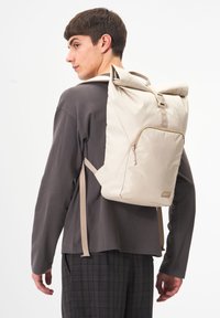 Cremfarbene Rolltop-Rucksack mit beigen Riemen, ausgestattet mit einer vorderen Reißverschlusstasche und einer glatten, matten Oberfläche. Verstellbarer Schnallenverschluss.
