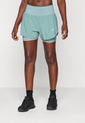 Personne portant un short de running superposé bleu clair, des chaussures de sport noires et des chaussettes noires, debout devant un fond uni.