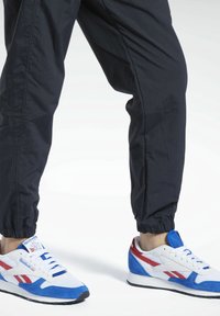 Valkoiset ja siniset urheilukengät punaisilla yksityiskohdilla, joissa on mokka- ja verkko-yläosa, nauhakiinnitys ja kumipohja. Mustat verkkarit on puettu.