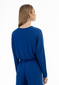 Indumento blu a maniche lunghe con vestibilità ampia, dotato di collo a giro e vita cinchata, realizzato in un tessuto liscio e elastico.