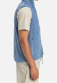 Světlomodrá fleecová vesta se zipovými kapsami a vysokým límcem, s měkkou texturou a bez rukávů.