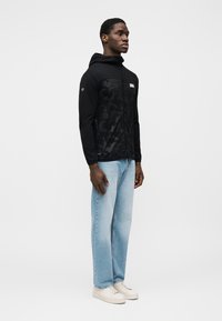 Μαύρο μπουφάν με κουκούλα και υφή, φερμουάρ μπροστά και λογότυπο σε πάτς. Συνδυάζεται με ανοιχτόχρωμο μπλε τζιν και μπεζ αθλητικά παπούτσια.