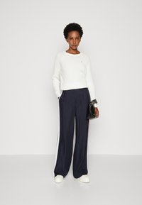 Maglione bianco a maglia, pantaloni wide-leg in satin blu navy con strisce bianche e rosse, e sneakers avorio; tiene una borsa nera.