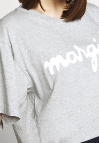En kvinna bär en grå oversized t-shirt med ordet "margi" i fet vit skrift, vilket visar en avslappnad och ledig stil.