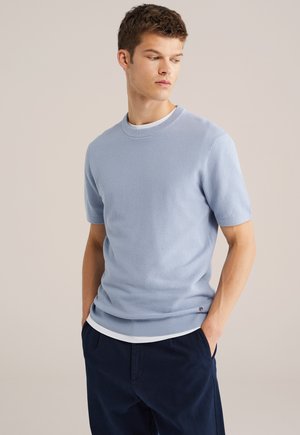 Jonge man die een lichtblauw gebreid kortärmelig trui draagt over een wit ondershirt met donkerblauwe broek, kijkend naar zijn linkerzijde.