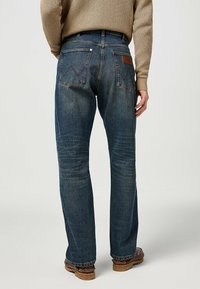 Jeans in denim blu scuro con taglio dritto, caratterizzati da due tasche posteriori con cuciture a contrasto, un'etichetta in pelle sulla vita e una texture invecchiata.