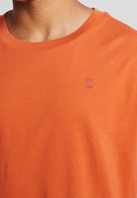 Oranžové bavlněné tričko s kulatým výstřihem. Na hrudi má jemně vyšitý logo v světlejším odstínu. Měkká textura.