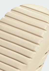 Sandale en caoutchouc beige à glissière avec une surface texturée, présentant des rainures horizontales et des points pour une meilleure adhérence. Bout arrondi avec un design minimaliste.