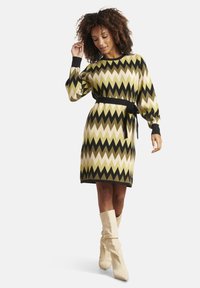 Vestito lavorato a maglia con motivo a zigzag in verde oliva, crema e nero; vita aderente con cintura; maniche lunghe; indossato con stivali beige.