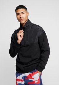 Felpa nera a pullover con design a mezza zip, caratterizzata da una parte superiore liscia nera in contrasto e una parte inferiore testurizzata.