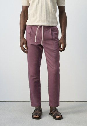 Hombre vistiendo pantalones de cordón color malva, camiseta de punto de manga corta color crema y sandalias de cuero marrón oscuro, de pie sobre un suelo gris.