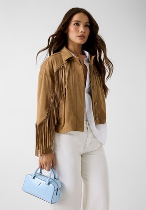 Femme portant une veste en suède marron à franges, chemise blanche, pantalon blanc, tenant un petit sac à main bleu clair sur un fond uni.