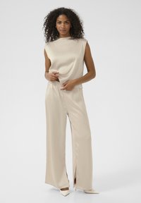Top sans manches beige avec un design drapé, associé à un pantalon large avec des fentes latérales. Le tissu a une texture soyeuse. Talons aiguilles.