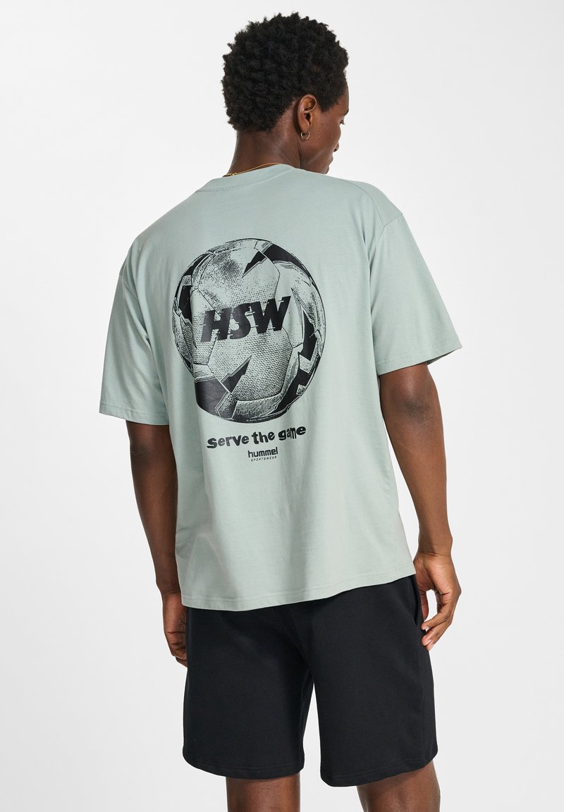 Mann trägt ein hellgrünes T-Shirt mit einem schwarzen grafischen Fußball und dem Text "HSW serve the game" auf dem Rücken, kombiniert mit schwarzen Shorts.