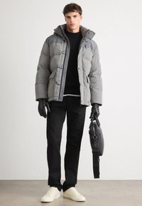 Chaqueta de plumas gris con capucha, cierre de botones en el frente y bolsillos laterales, usada sobre un suéter negro, combinada con pantalones negros y zapatillas blancas.