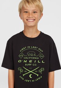 Camiseta negra con un estampado gráfico verde de un oso, tablas de surf cruzadas y palmeras. Manga corta con cuello redondo.