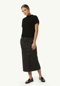 Schwarzer Kurzarm-Pullover mit subtiler Textur, kombiniert mit einem langen schwarzen knöpfbaren Rock. Das Model trägt schwarze Slipper und steht im Profil.