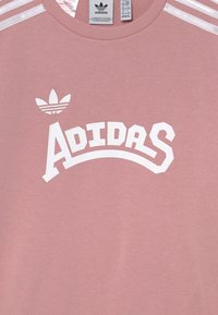 Rosa Adidas sweatshirt med vit logotext och trefoil-symbol, med vita ränder på axelsömmarna och synliga storleks- och skötselinstruktioner.
