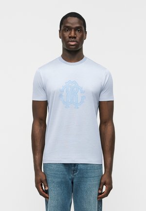Homme portant un t-shirt bleu clair à col rond avec un monogramme décoratif subtil sur la poitrine et un jean bleu délavé moyen, sur fond blanc.