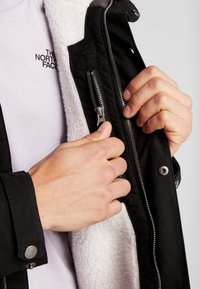 Les mains d'une personne qui ferment une veste noire doublée de polaire blanche sur une chemise blanche avec un logo texte noir sur la poitrine.