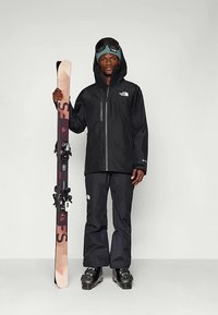 Fekete sídzseki és nadrág logós részletekkel, síléceket tartva. Kapucnival, hó-szemüveggel és fekete síbakancsokkal egy semleges háttér előtt.