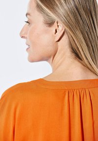 Leichte orange Bluse mit weitem Ausschnitt und plissiertem Rückendetail, aus glattem Stoff mit entspanntem Schnitt.