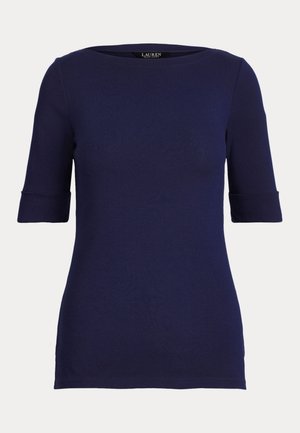 Une femme présente dans un studio un haut bleu marine, un pantalon blanc raccourci à jambes larges, des sandales marron et un petit sac à main marron.