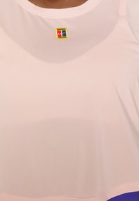 Nahaufnahme eines blassrosa Kleidungsstücks mit einem kleinen, quadratischen, bestickten Logo in Rot, Marineblau, Gelb und Weiß in der Nähe des Halsausschnitts.