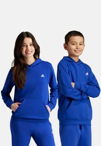 Felpe con cappuccio blu realizzate in materiale morbido, con tasche frontali, polsini a coste e un piccolo logo bianco. Indossate da una ragazza e un ragazzo.