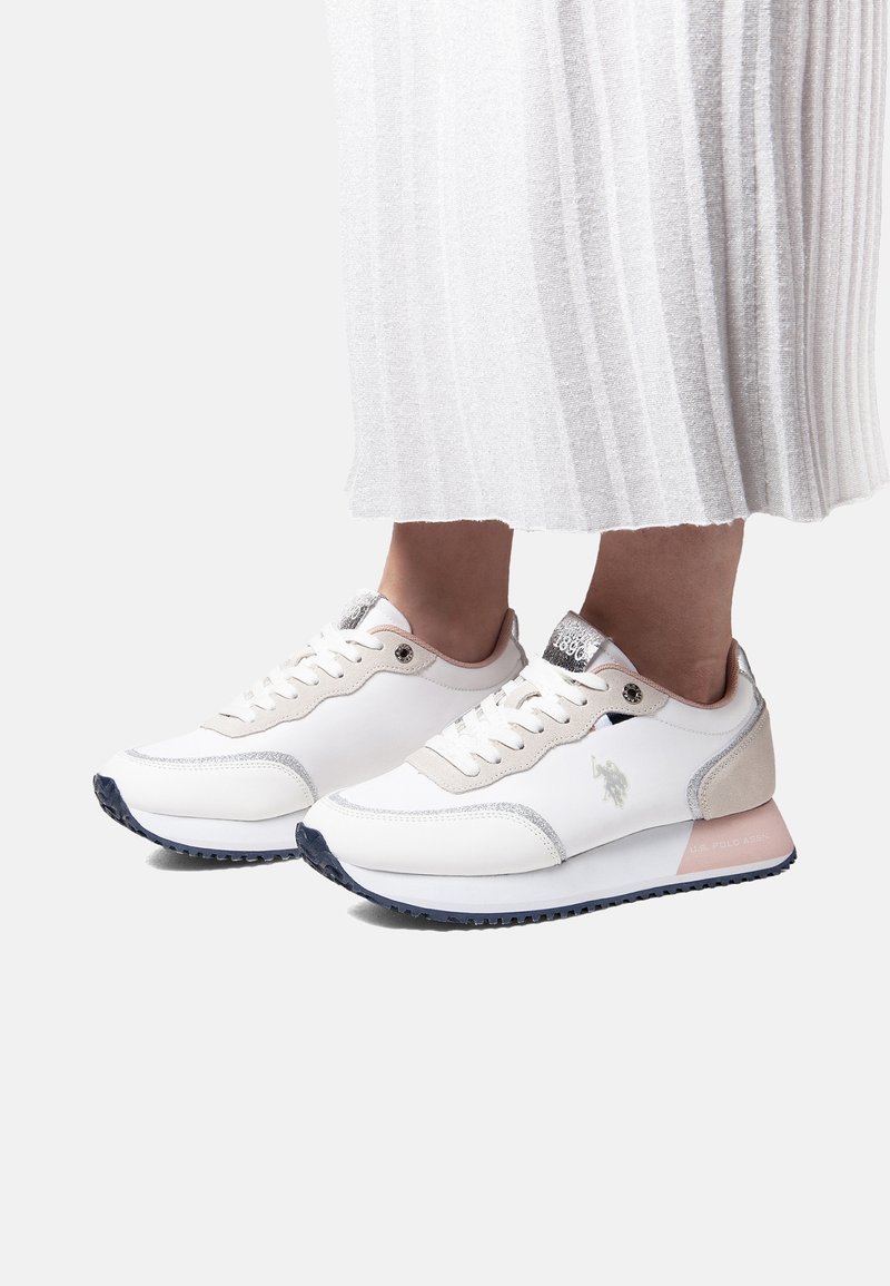 Hvide og beige snøresneakers med sorte såler, båret med en hvid plisseret nederdel, vist på en lys baggrund.