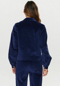 Temno modra kordurova jakna s razširjenim ovratnikom, dolgimi nabranimi rokavi in gumbnimi manšetami. Gladka tekstura s skrajšanim dizajnom, v kombinaciji s usklajenimi hlačami.