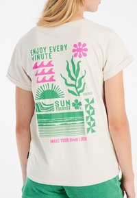 Camiseta beige con estampados gráficos coloridos en verde y rosa, con frases y patrones en la parte posterior, mangas cortas enrolladas y un corte relajado.