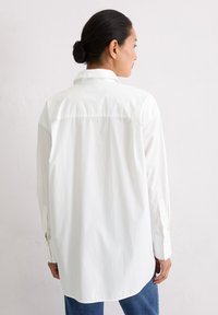 Frau mit dunklem Dutt trägt ein lockeres weißes Langarmshirt und blaue Jeans und steht vor einer schlichten weißen Wand.