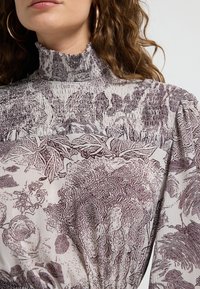 Tekstilkjole i let stof med en høj flæsekrave. Mønstret med brune blomsterdesigns, samlet ved midjen, med pufærmer.