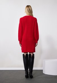 Robe tricotée rouge à manches longues, avec des poignets côtelés et une coupe décontractée, associée à de hautes bottes en cuir noires avec un design découpé.