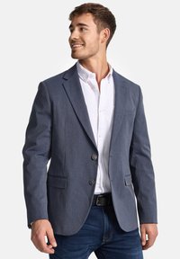 Dark blue blazer made of textured fabric, featuring two buttons, two front pockets, and tailored fit, worn over a white shirt.