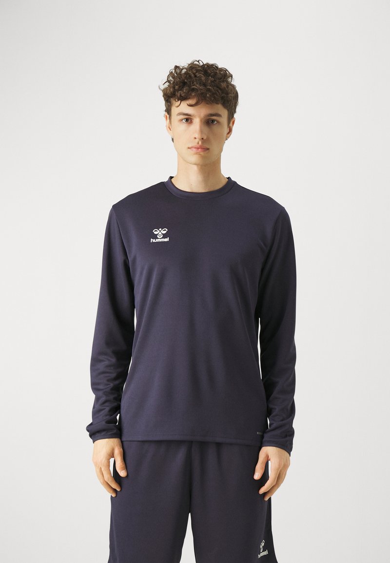 Jeune homme aux cheveux bouclés portant un t-shirt de sport Hummel à manches longues foncé et un pantalon assorti, debout devant un fond blanc uni.